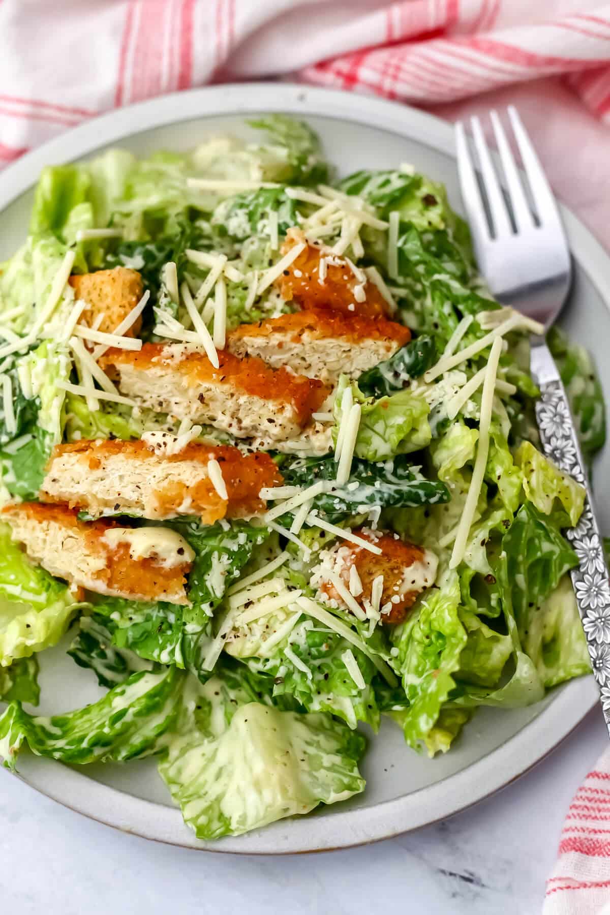 A white plate filled with romaine lettuce, vegan chicken strips, with vegan Caesar dressing, and parmesan cheese sprinkled on top.