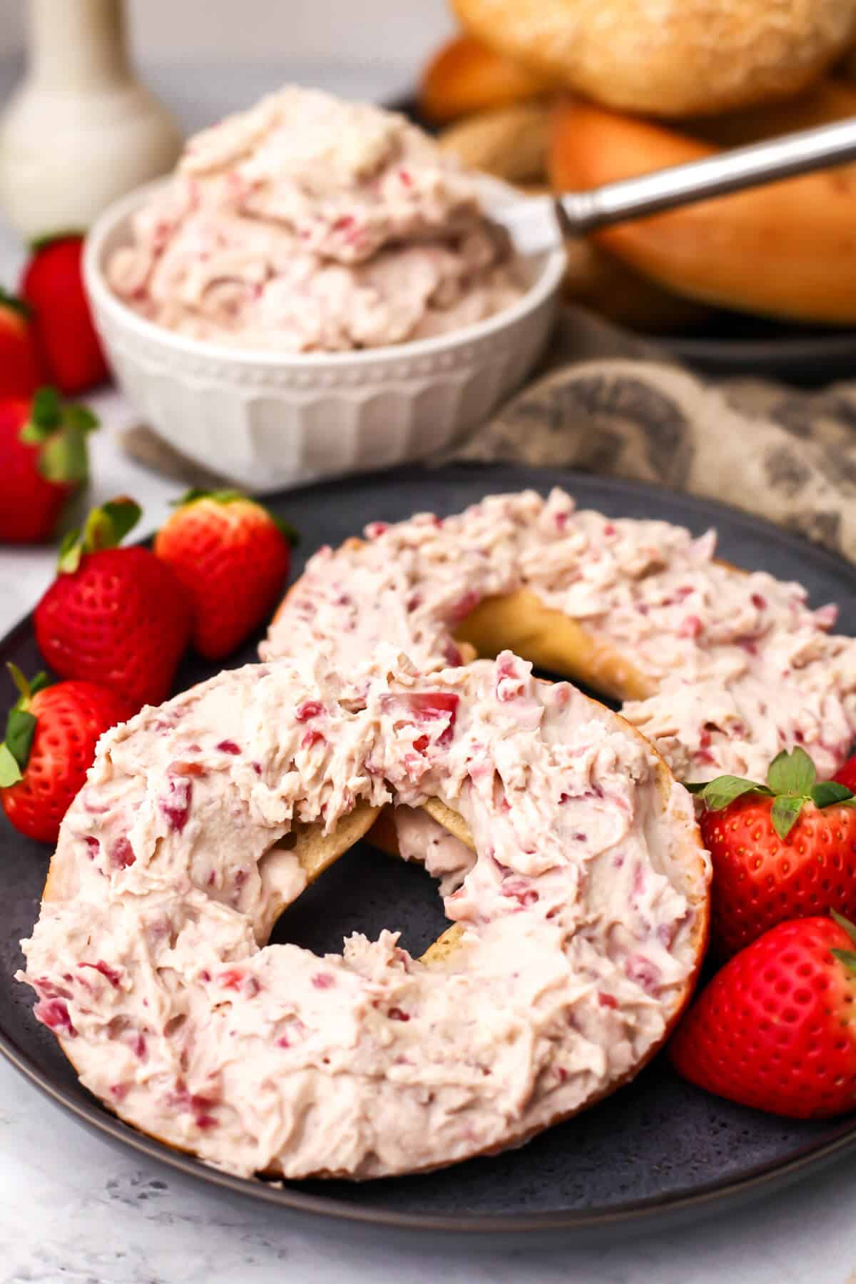 A bagel with vegan strawberry cream cheese and fresh strawberries on the side with more bagels behind it.