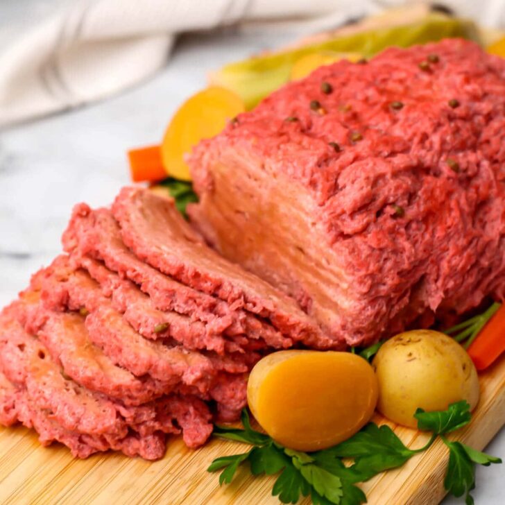 A seitan corned beef roast sliced on a cutting board with potatoes, carrots, and cabbage around it.