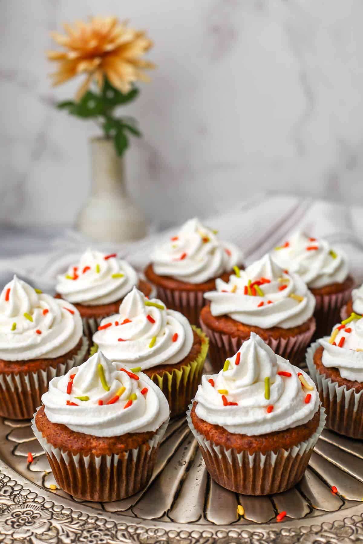 A platter full of vegan carrot cake muffins with cream cheese frosting and orange and green sprinkles with an orange flour behind them.