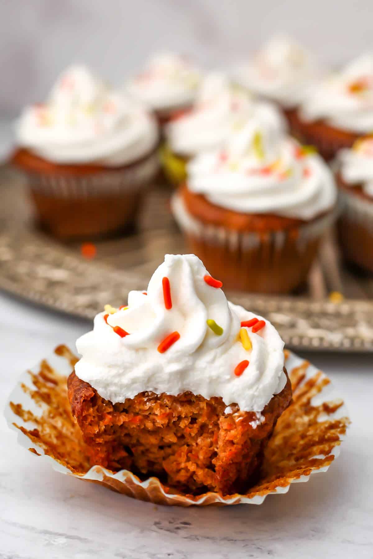 A vegan carrot cupcake with a bite out of it with a plate of cupcakes behind it.