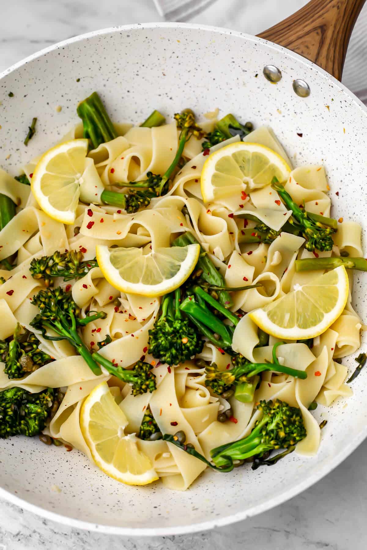 Vegan broccolini pasta with garlic, lemon, and capers in a white wok with lemon slices on it.