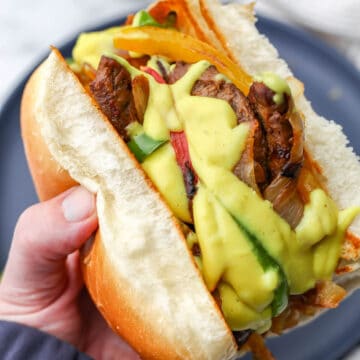 A vegan Philly cheese steak with grilled onions, peppers, seitan, and vegan cheese sauce being held in someone's hand.
