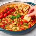 A blue cast iron pan filled with butter beans in a tomato pesto sauce with someone dipping toast in it.