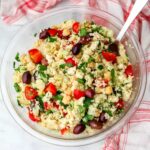 A top view of a vegan couscous salad with tomatoes, cucumbers, olives, and artichokes with a red and white tea towel.
