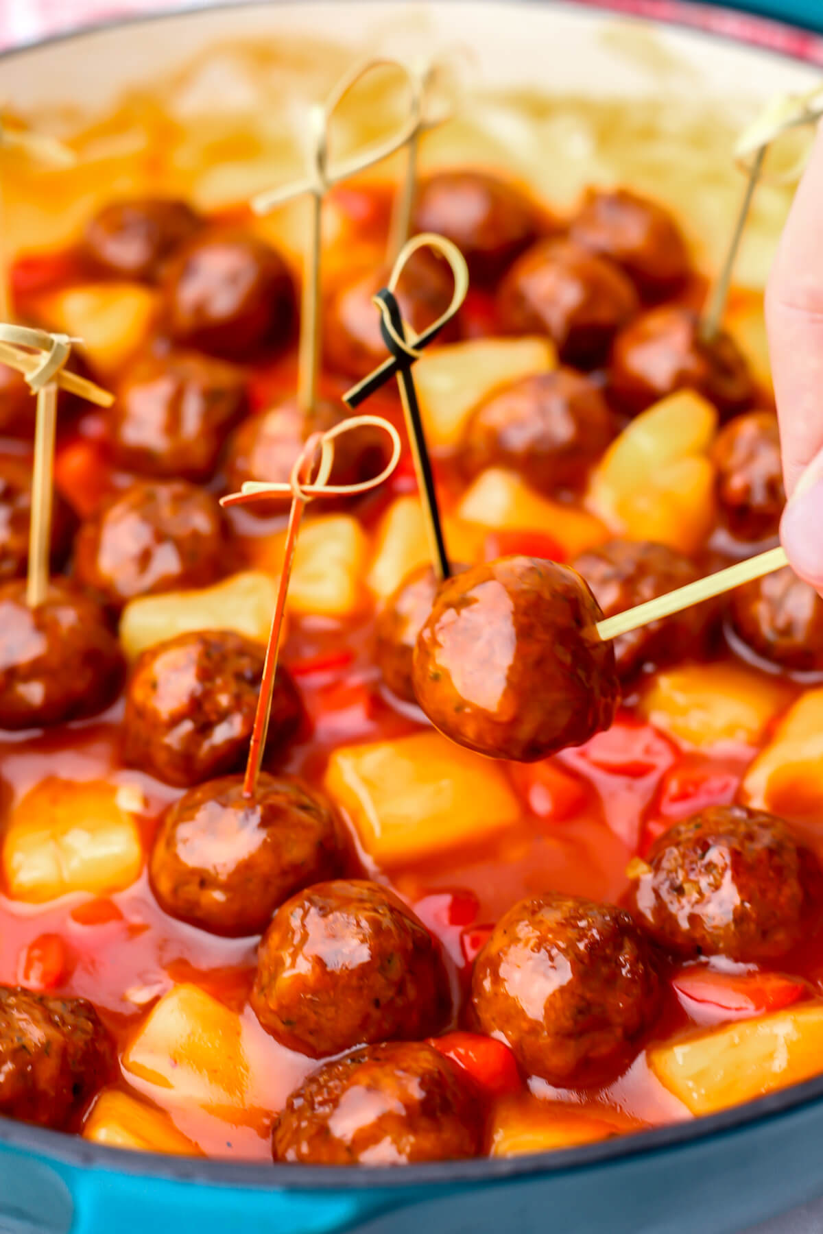 Someone picking up a tooth pick with a vegan sweet and sour meatball out of a pan of vegan meatballs.