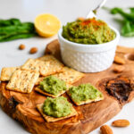 A bowl of almond pate made with fresh basil, lemon juice and spices in a small white bowl with some spread on crackers in front of it.