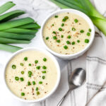 Two bowls of creamy vegan cauliflower potato leek soup with leeks on the side.