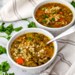 Two bowls of vegan beef barley soup with parsley and a tea towel around it.