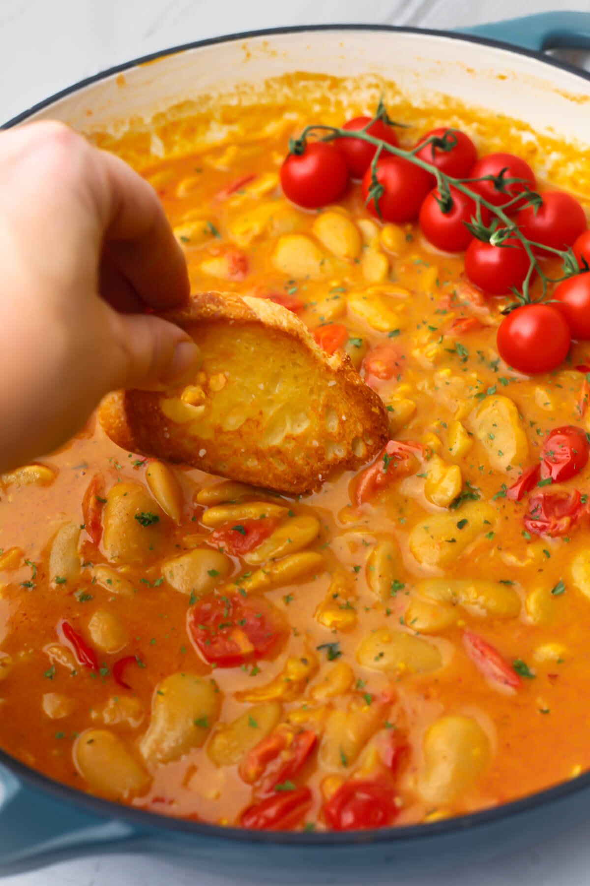 Crispy bread being dipped into vegan curried butter beans in a large blue skillet.