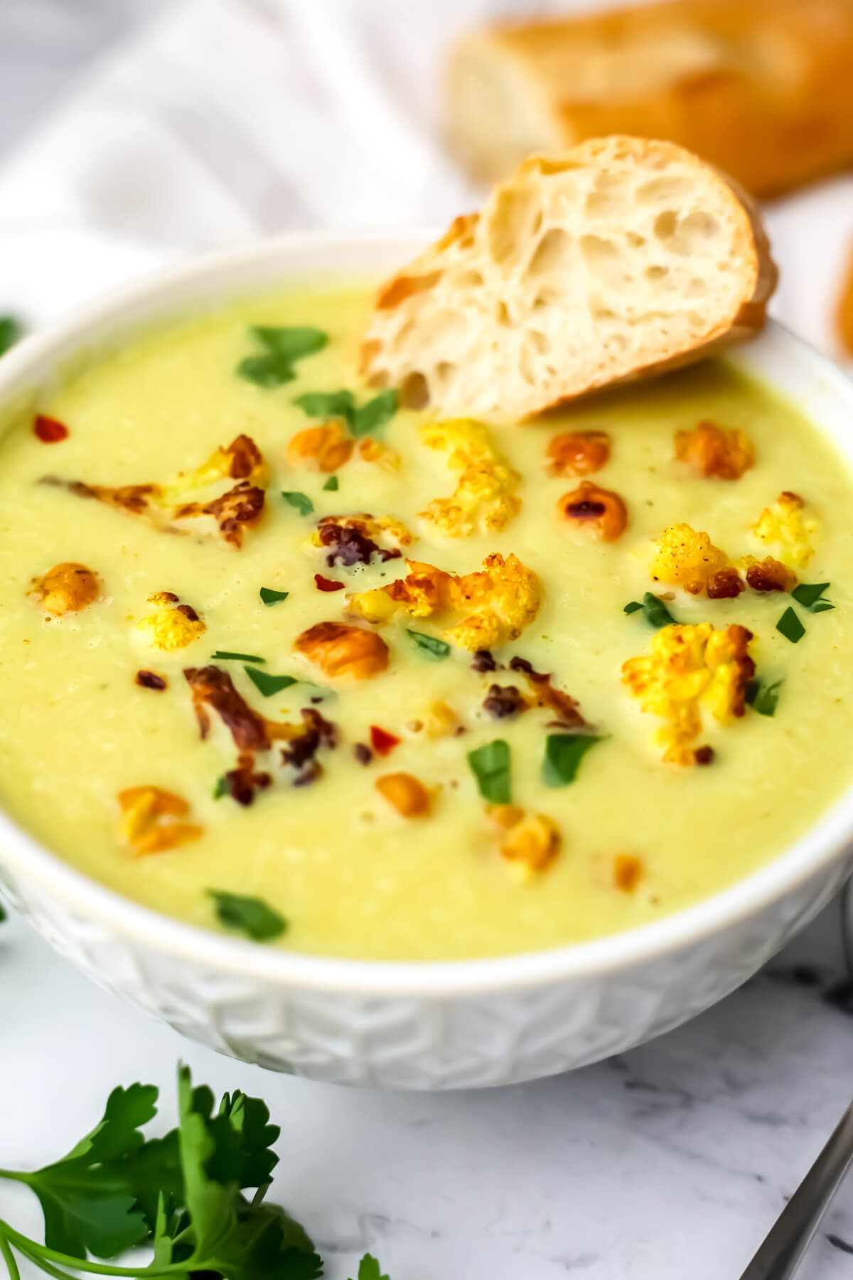 A white bowl filled with vegan curried cauliflower soup with roasted cauliflower and chickpeas on top and a piece of bread on the side.