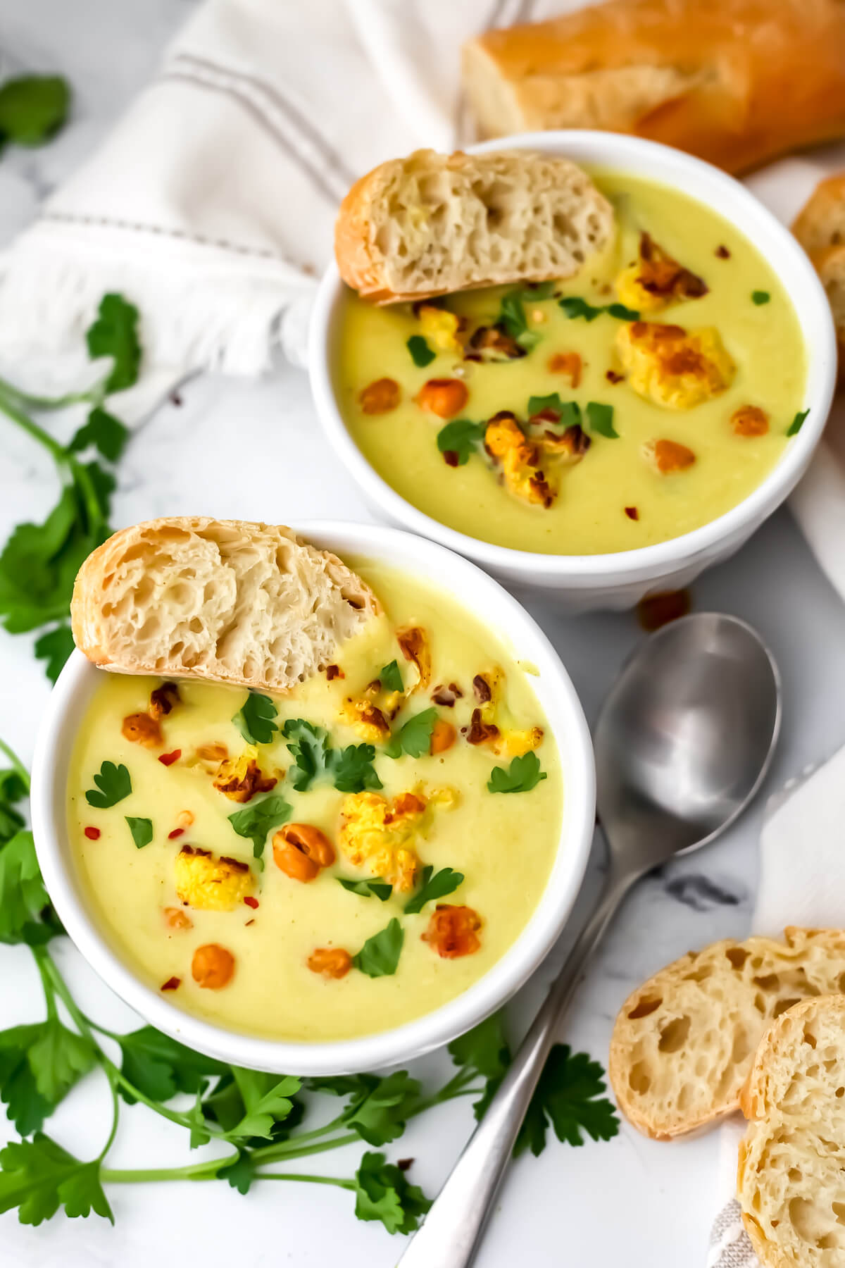 A top view of 2 bowl of vegan curried cauliflower and chickpea soup with bread and parsley on the side.