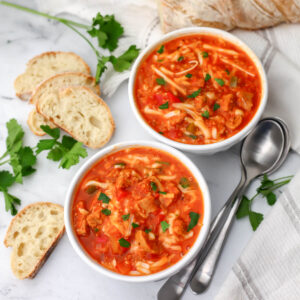 A top view of two bowl of vegan stuffed pepper soup topped with cheese and parsley and with bread and spoons on the side.