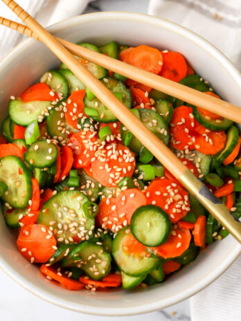 A top view of a carrot and cucumber salad in a white bowl with chopsticks on top.