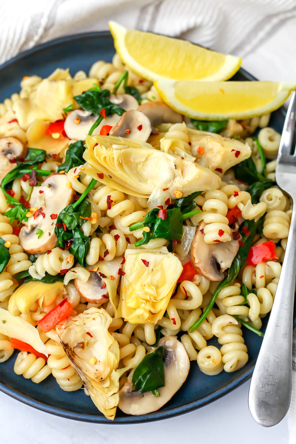A top view of a blue plate filled with pasta topped with artichokes, bell peppers, mushroom and spinach with lemon wedges on the side.