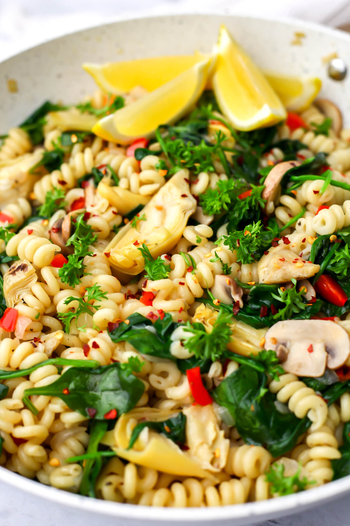A large white skillet filled with pasta and a sauce made from marinated artichokes, onions, peppers, mushrooms, and spinach with lemon wedges on the side.
