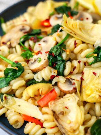 A plate full of vegan artichoke pasta with red bell peppers, mushrooms, and spinach.