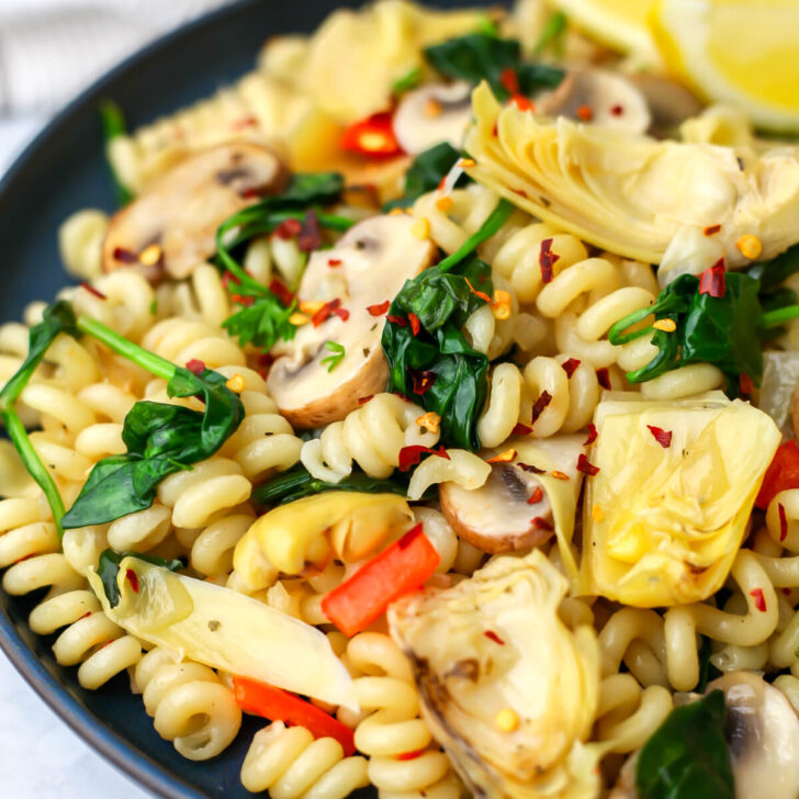 A plate full of vegan artichoke pasta with red bell peppers, mushrooms, and spinach.
