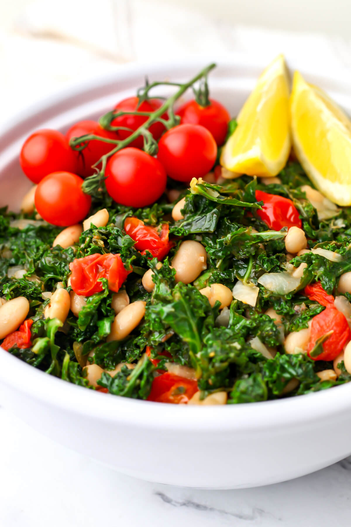 A vegan kale white bean salad with tomatoes and lemon wedges on the side.