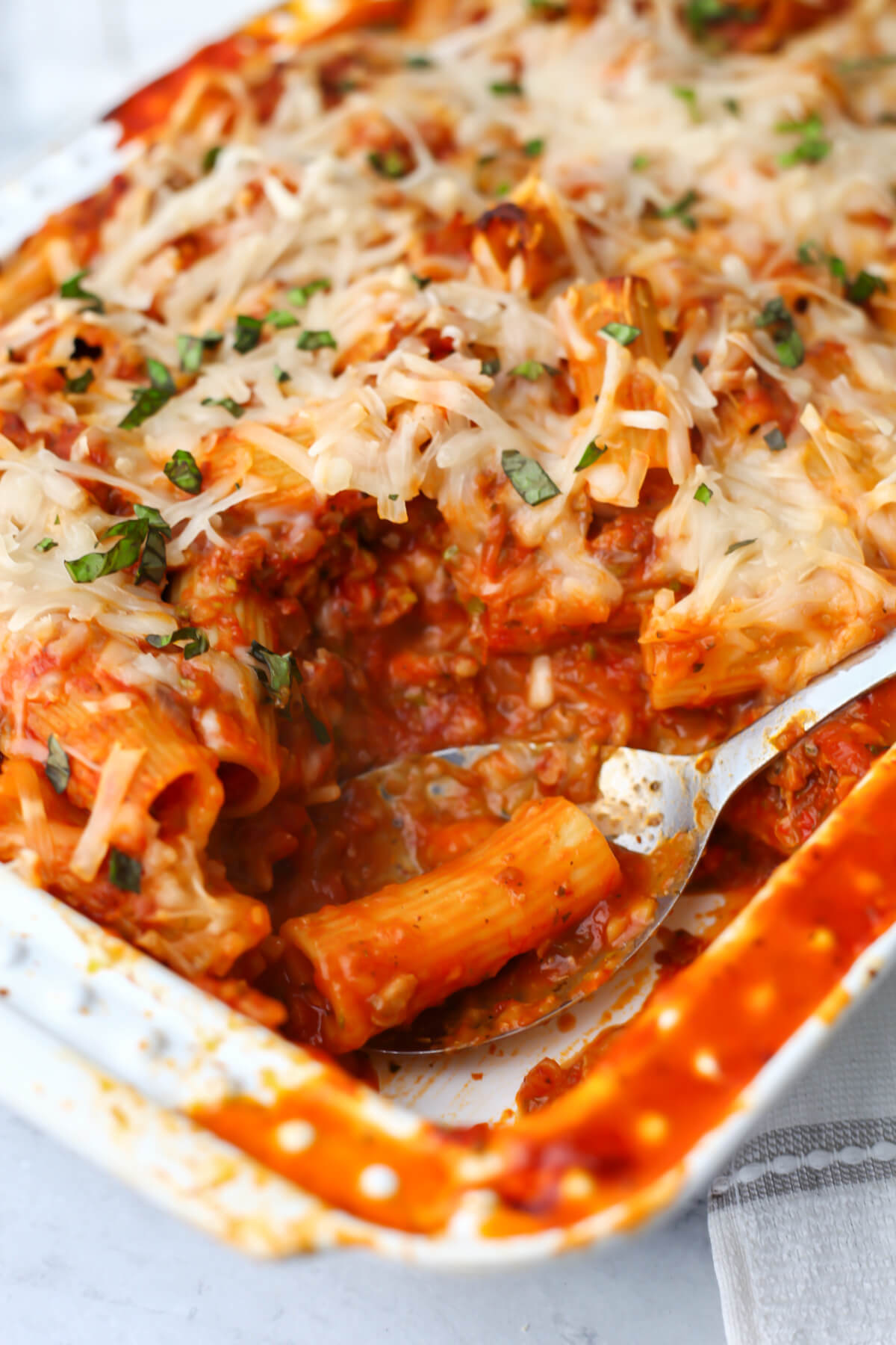 A baking dish filled with vegan rigatoni pasta baked with vegan cheese on top with a serving spoon in it.