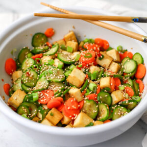 A white bowl filled with cucumber tofu salad with carrots, green onions, and sesame seeds.
