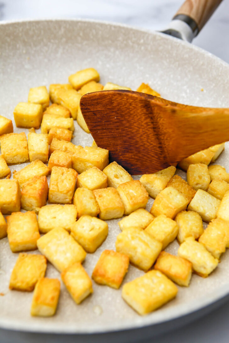 Tofu cubes frying in a pan being stirred with a wooden spoon.