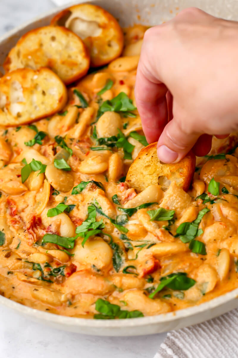 A frying pan filled with Marry Me Butter Beans being scooped up with a slice of toasted bread.
