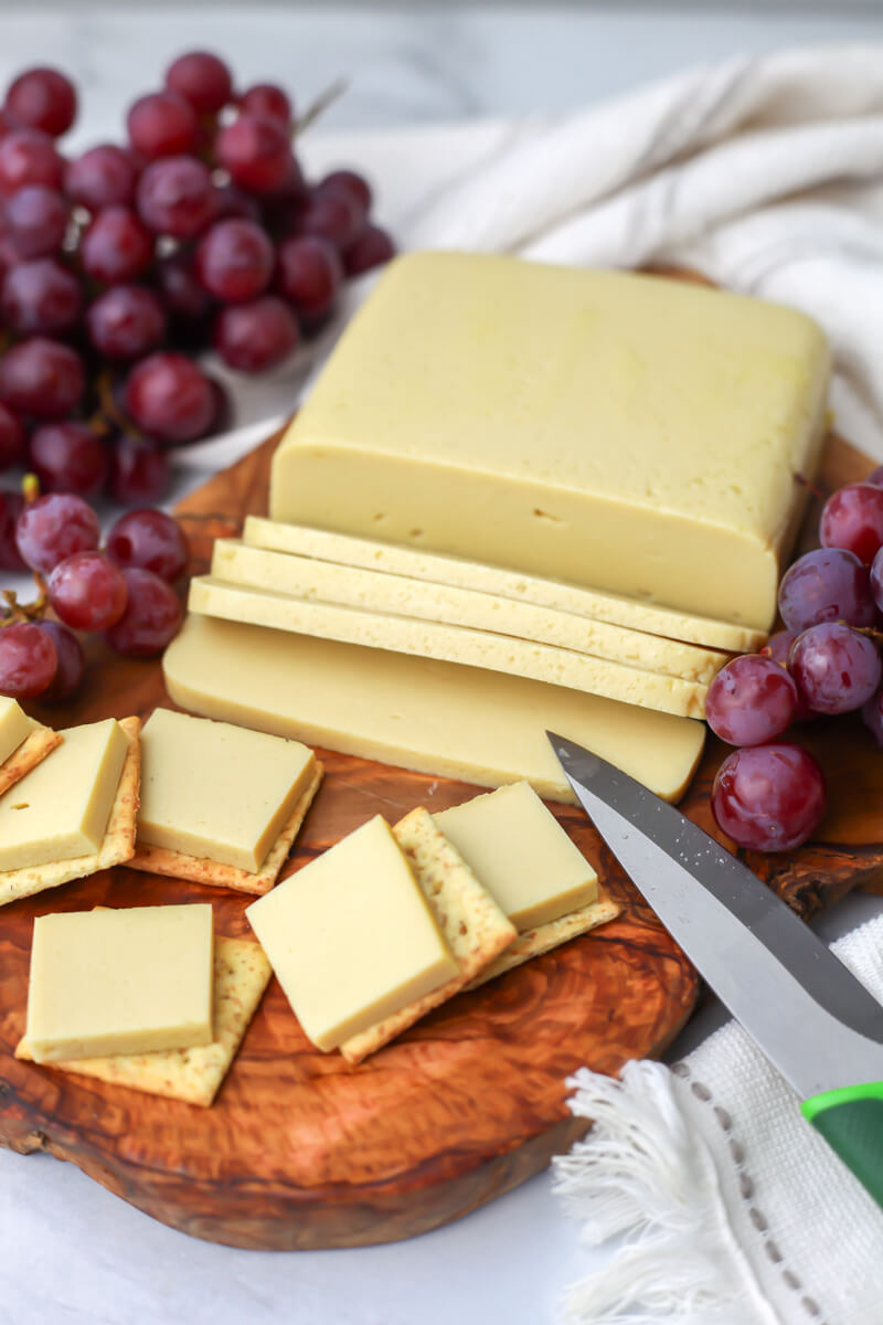 Vegan cheese made from silken tofu sliced on a wooden cutting board with grapes on the side.