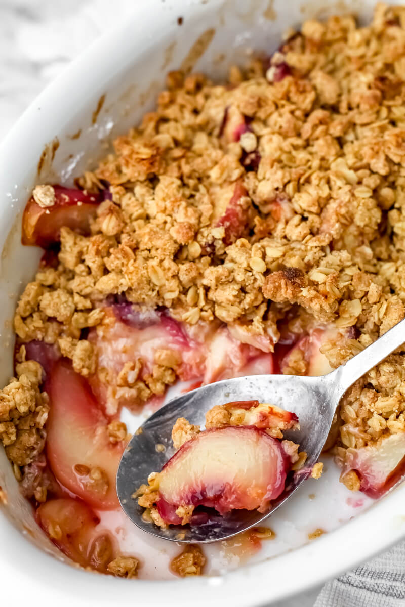 A baking dish full of vegan peach crisp with a scoop taken out of it and a peach slice on a spoon.