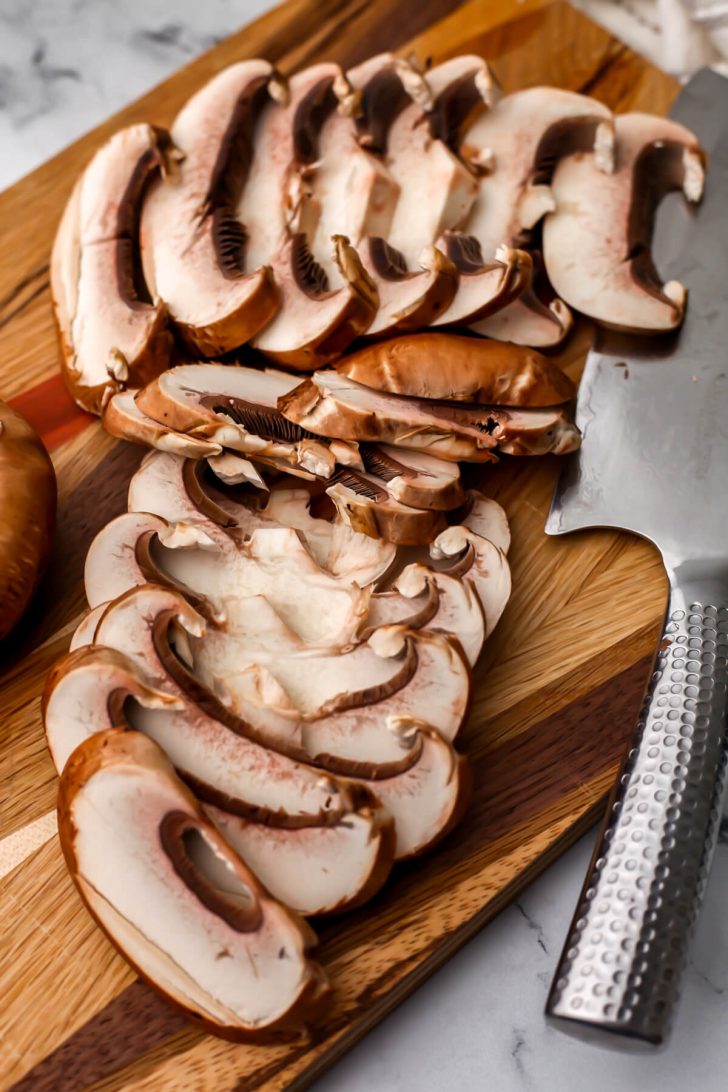 Slice portobello mushrooms on a wooden cutting board with a knife on the side.
