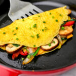 A Just Egg omelet filled with veggies in a red iron skillet being picked up with a spatula.