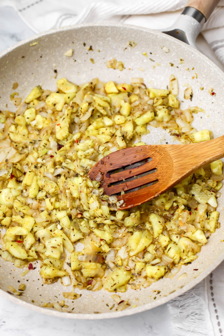 Onions, garlic, and chopped zucchini sauteing in a pan being stirred with a wooden spoon.
