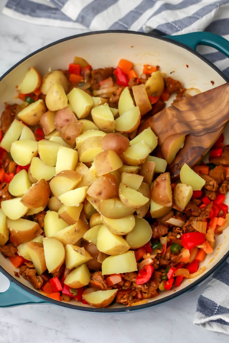 Cooked potato cubes being added to onions, peppers and vegan sausage in a skillet to make vegan hash.