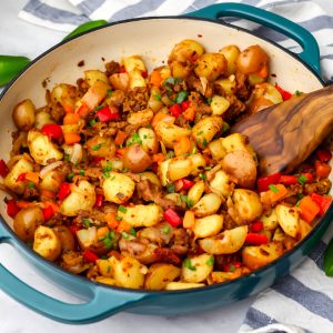 Vegan breakfast sausage made with onions, peppers, potatoes, and vegan sausage in a blue skillet being stirred with a wooden spoon.