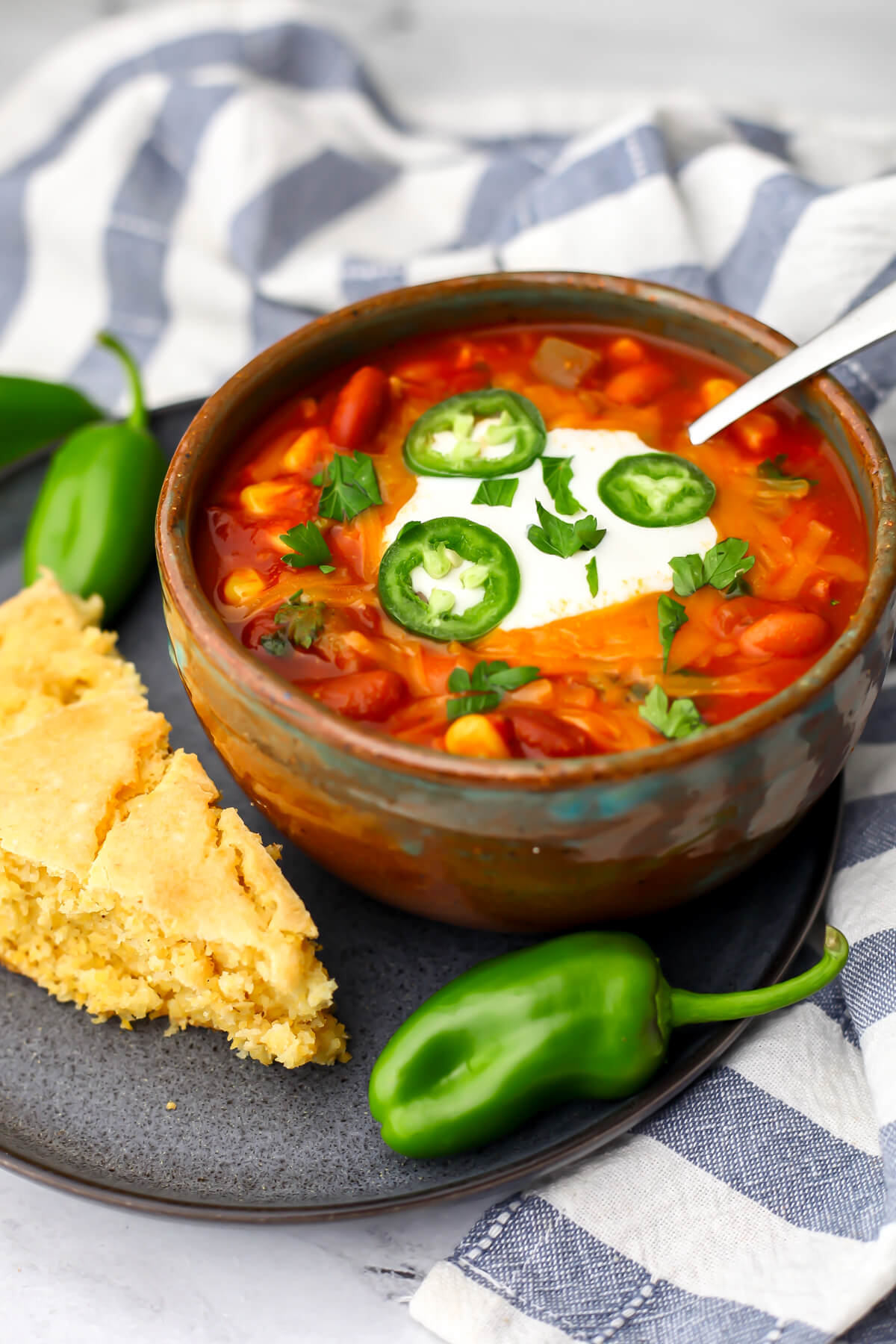 A bowl of vegan chili on a plate with a slice of corn bread and jalapenos.