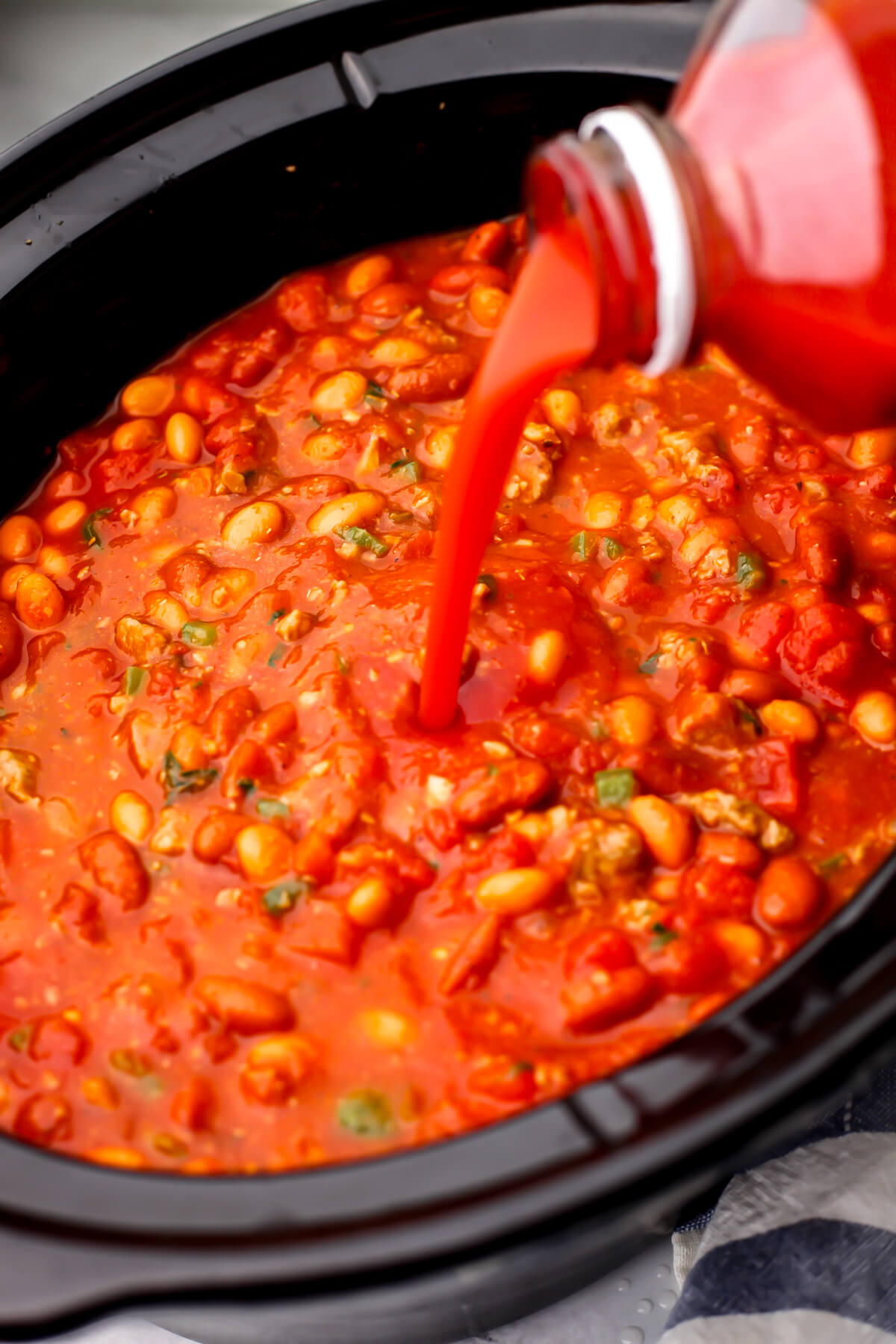 Tomato juice being added to vegan chili in a slow cooker.
