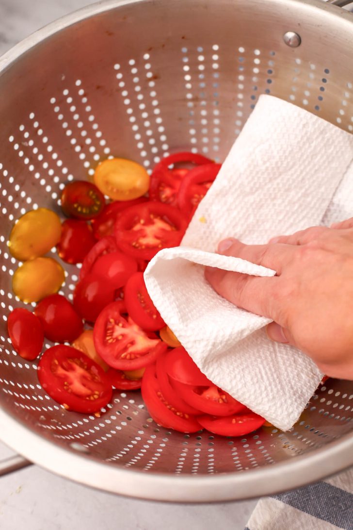 Sliced tomatoes in a colander sprinkled with salt being patted with a paper towel to get off excess liquid.