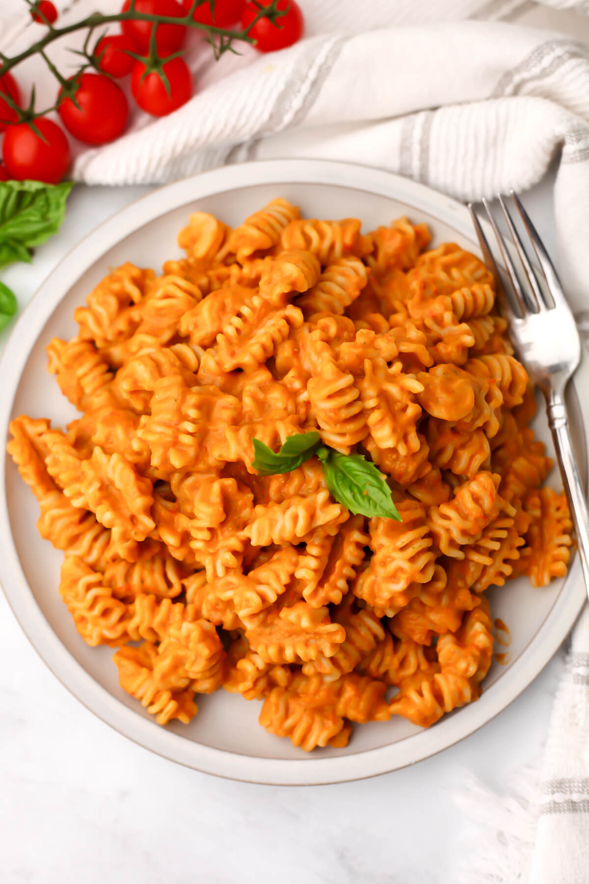 A top view of a creamy roasted vegetable pasta with a leaf of basil on top and cherry tomatoes next to it.