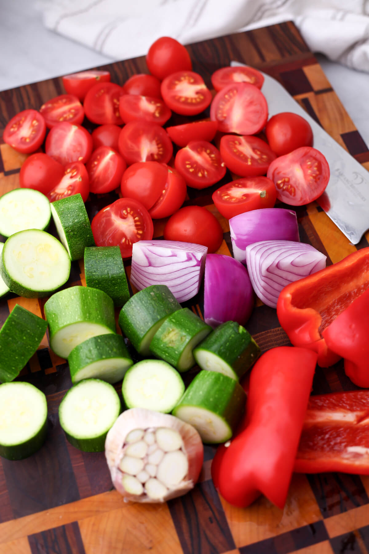 Tomatoes, red bell peppers, zucchini, red onion, and a head of garlic on a cutting board.