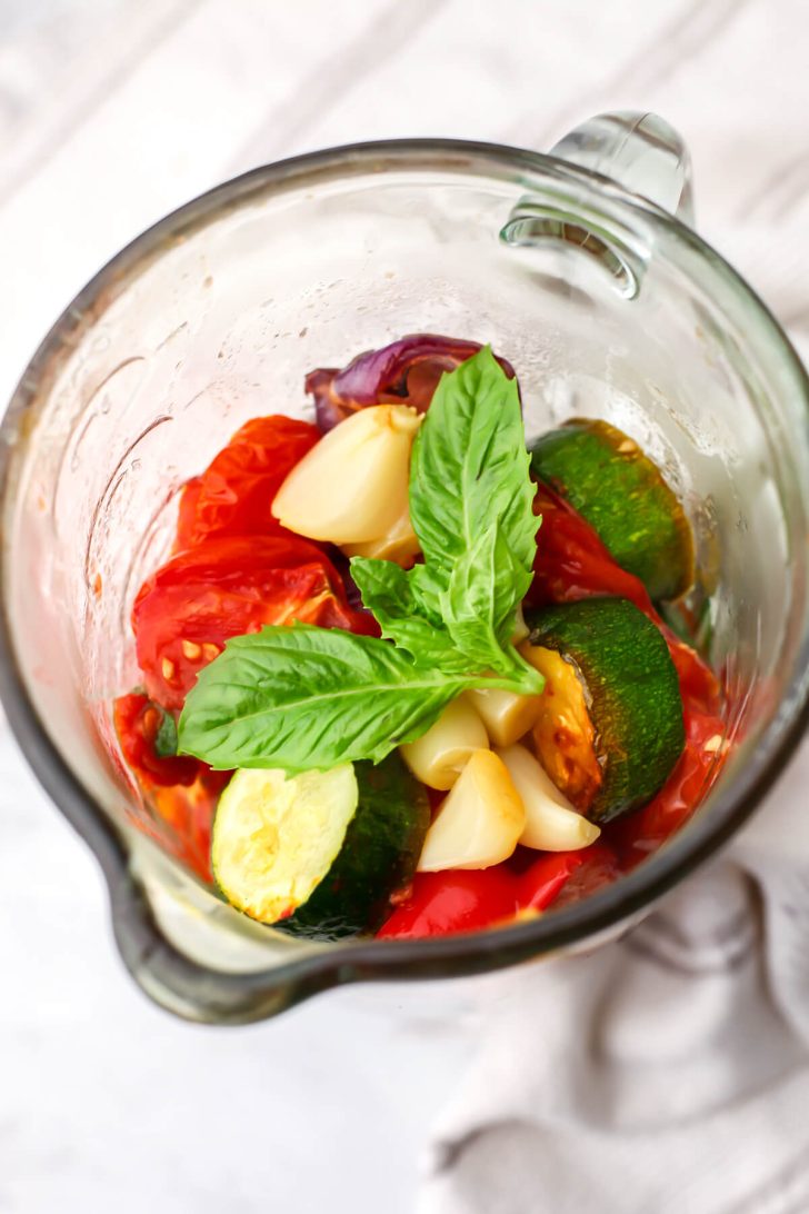 Roasted vegetables added to a blender with olive oil and fresh basil.