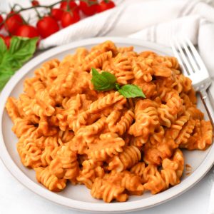 A white plate filled with roasted vegetable pasta sauce mixed with pasta with a fork on the side and cherry tomatoes behind it.