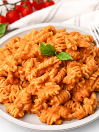 A white plate filled with roasted vegetable pasta sauce mixed with pasta with a fork on the side and cherry tomatoes behind it.