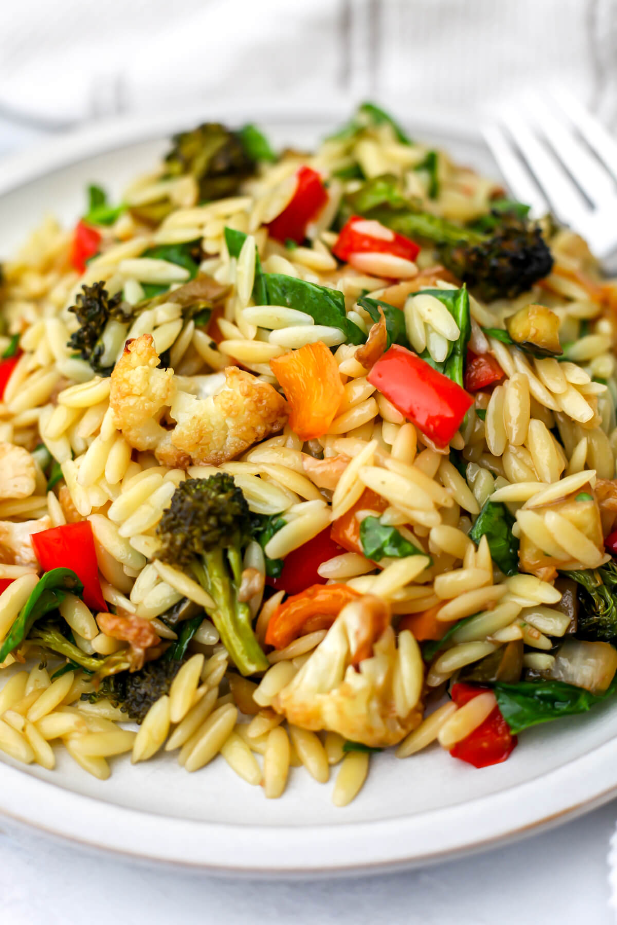 A white plate full or roasted vegetable orzo salad with a fork on the side.
