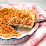 A slice of a vegan Dutch apple pie with a crumble topping being taken out of a pie dish.