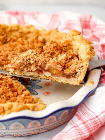 A slice of a vegan Dutch apple pie with a crumble topping being taken out of a pie dish.