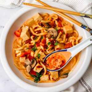 A white bowl full of noodles and tofu in a Thai red curry coconut sauce.