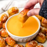 A soft pretzel bit being dipped into a bowl of vegan beer cheese sauce.
