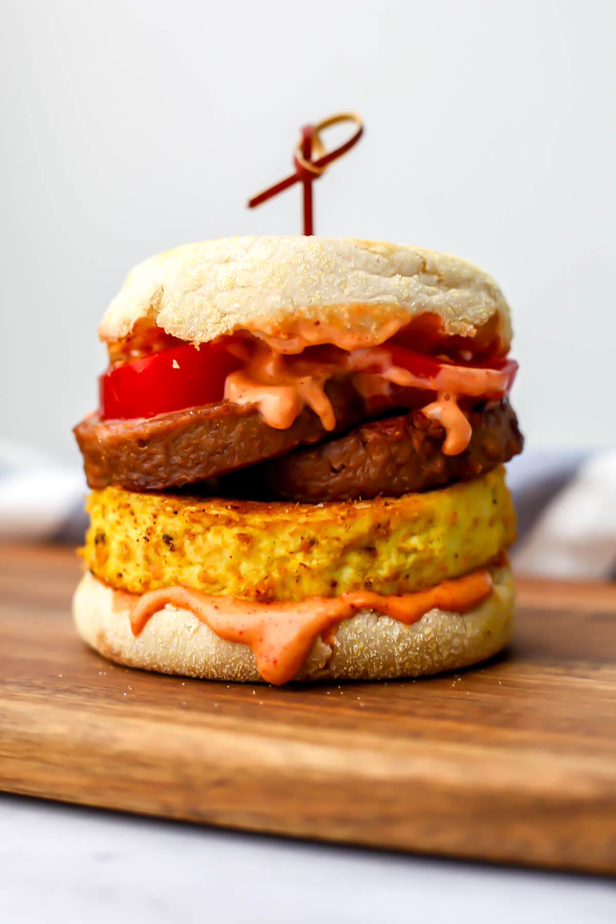 A tofu breakfast sandwich on an English muffin with tomatoes vegan sausage, and a creamy sauce sitting on a cutting board.