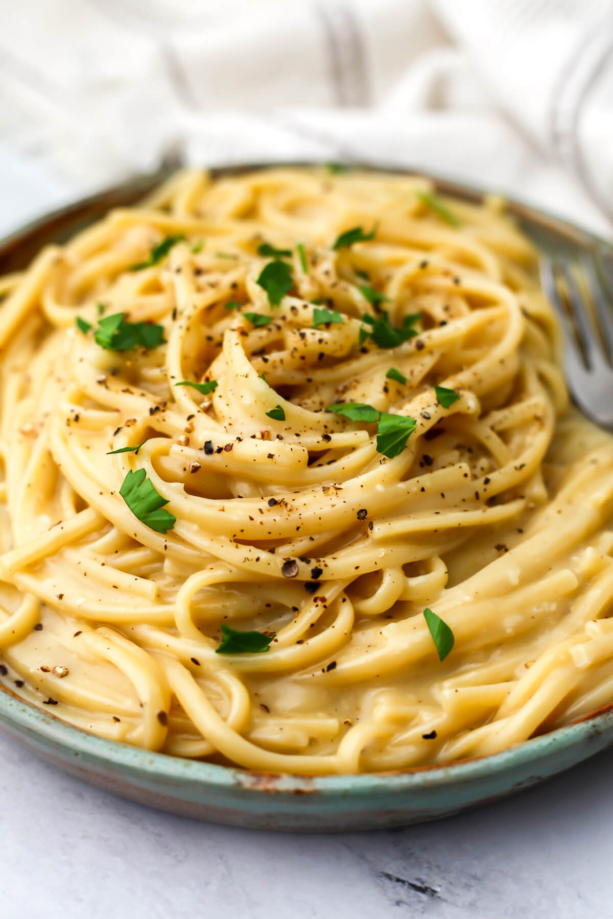 A plate of vegan cacio e pepe pasta topped with black pepper and parsley on a green plate with a fork on the side.