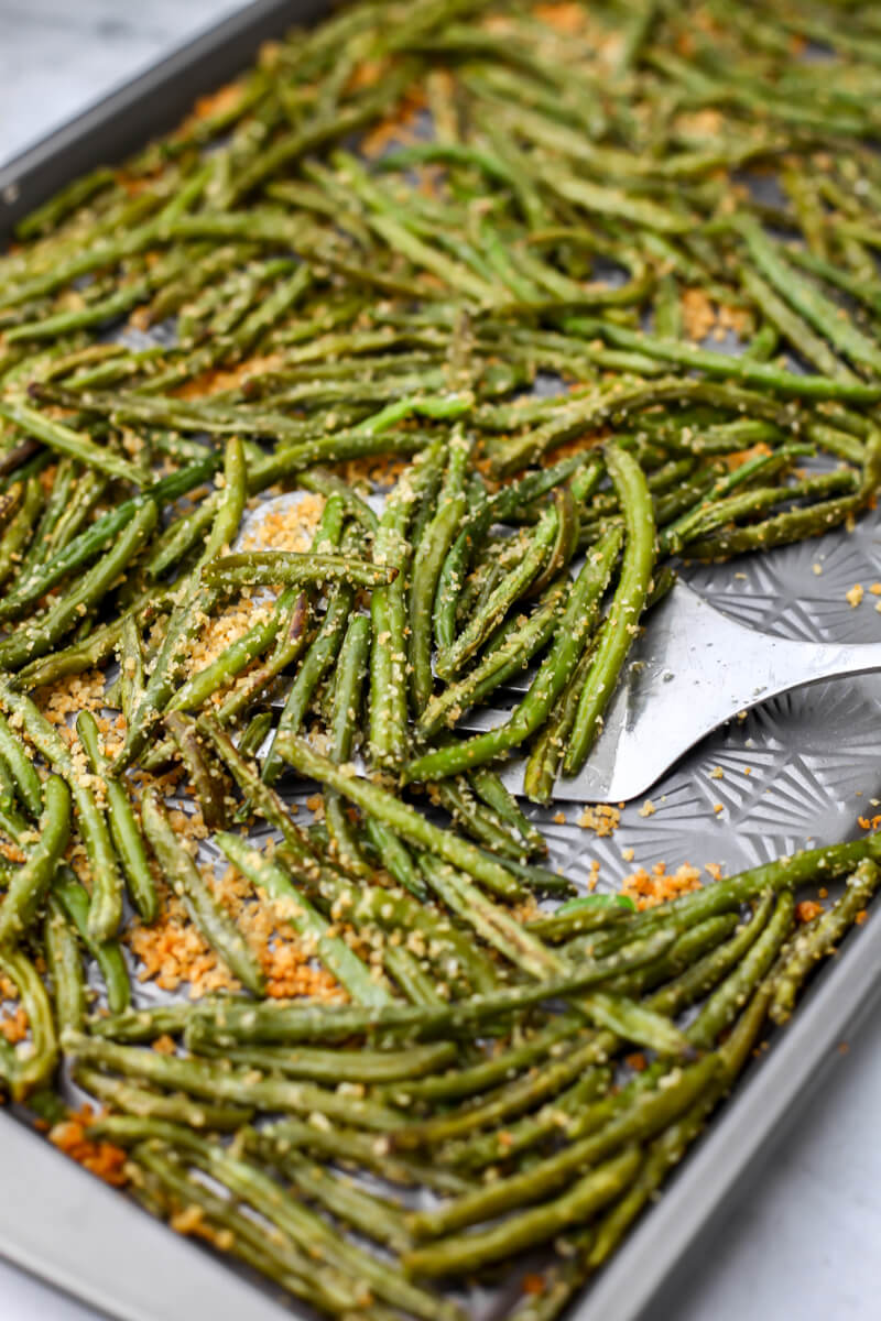 A baking sheet with roasted green beans coated with garlic breadcrumbs being scooped up with a spatula.